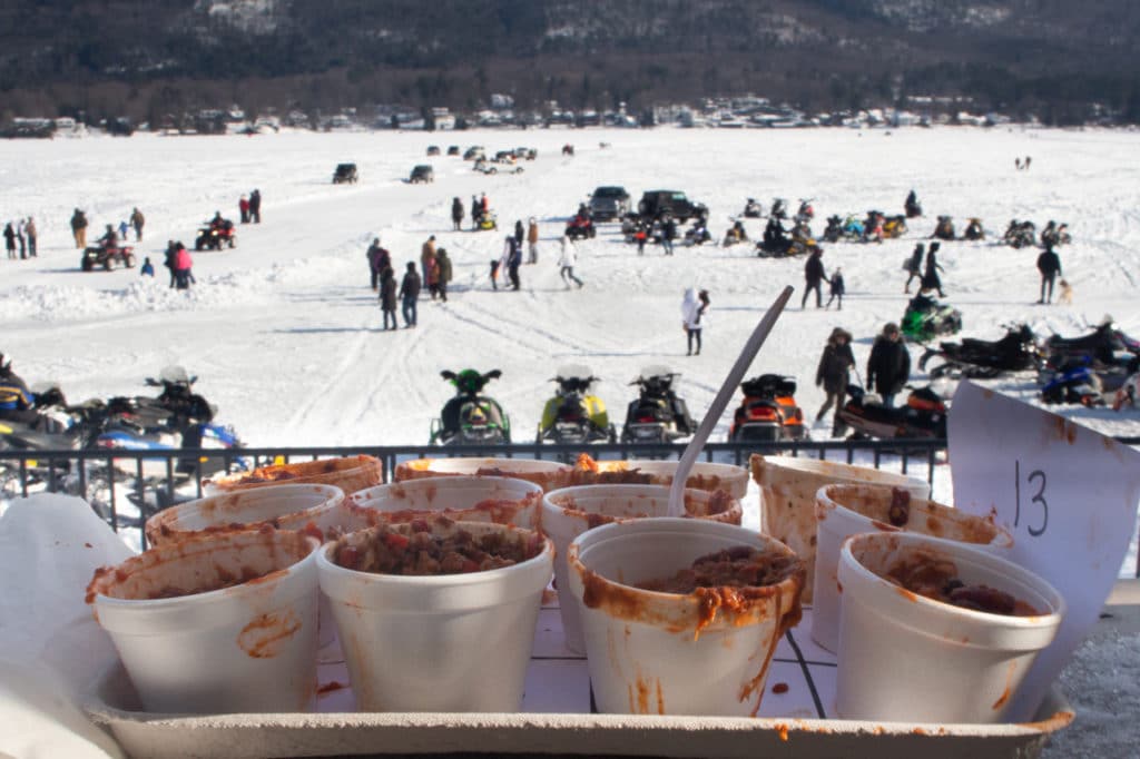 A tray of small styrofoam bowls full of chili, overlooking a frozen lake crowded with several people. 