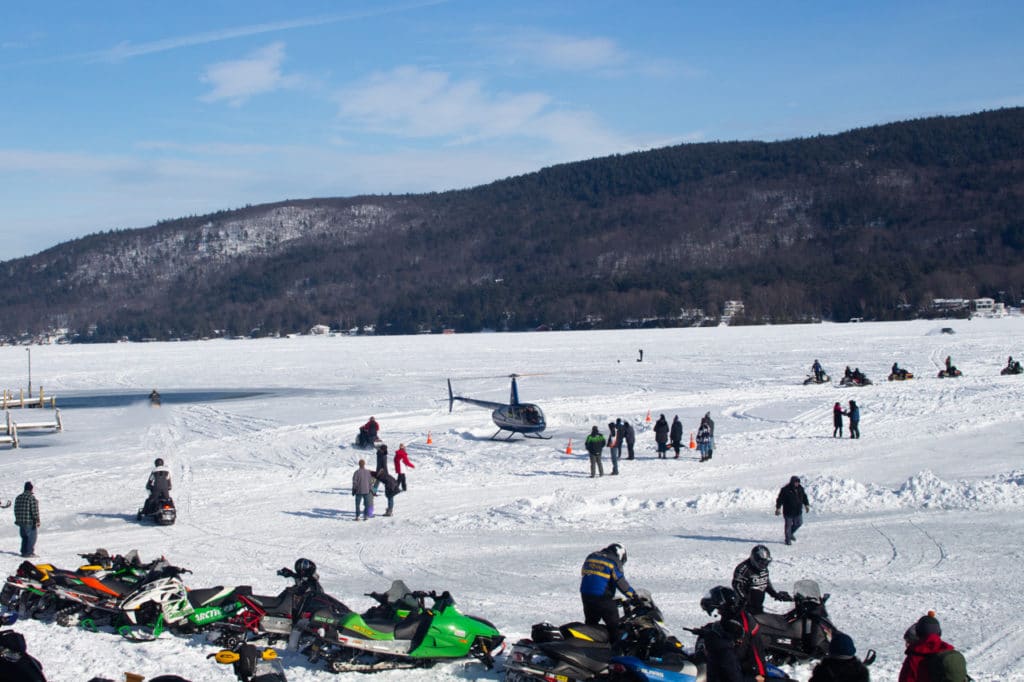 Snowmobiles and a helicopter parked on a frozen lake. Several people are milling around. 