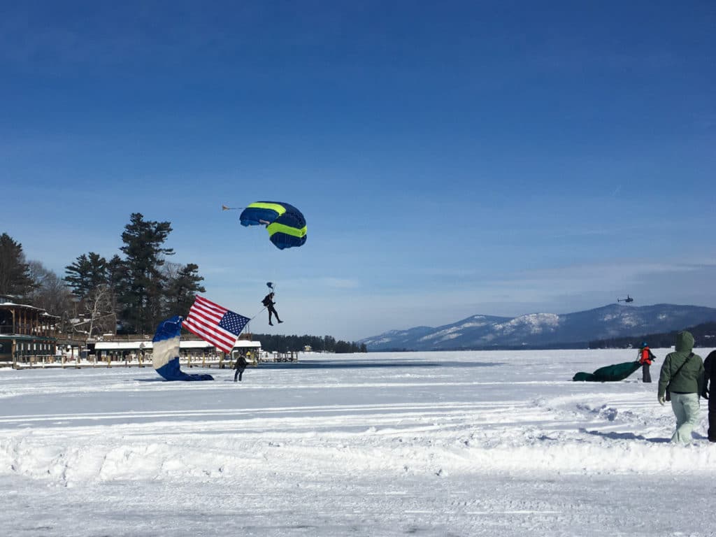 A man landing a parachute, holding a U.S. flag, onto a frozen lake. 