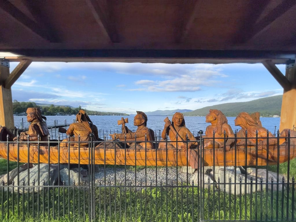 Large carved wood monument depicting six men rowing a canoe. Lake George is in the background.
