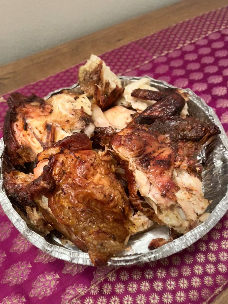 A whole grilled chicken chopped into quarters inside an aluminum takeout container. 