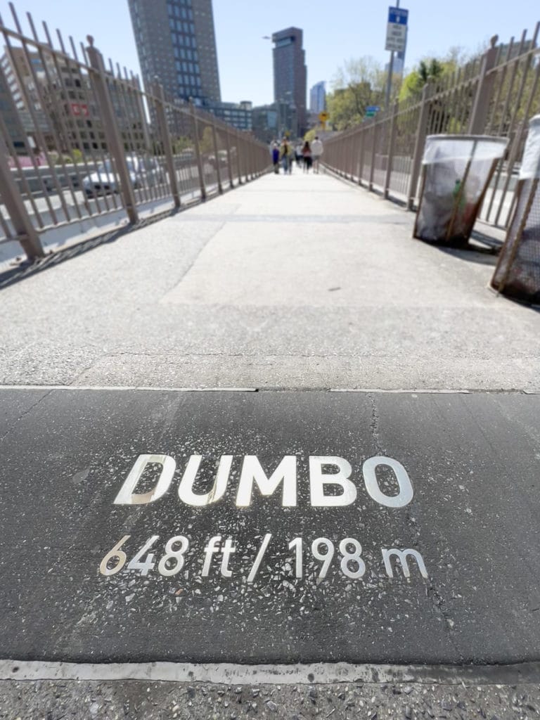 A sign embedded into the concrete ground that says DUMBO 648 ft/198 m. At the Brooklyn end of the Brooklyn Bridge.