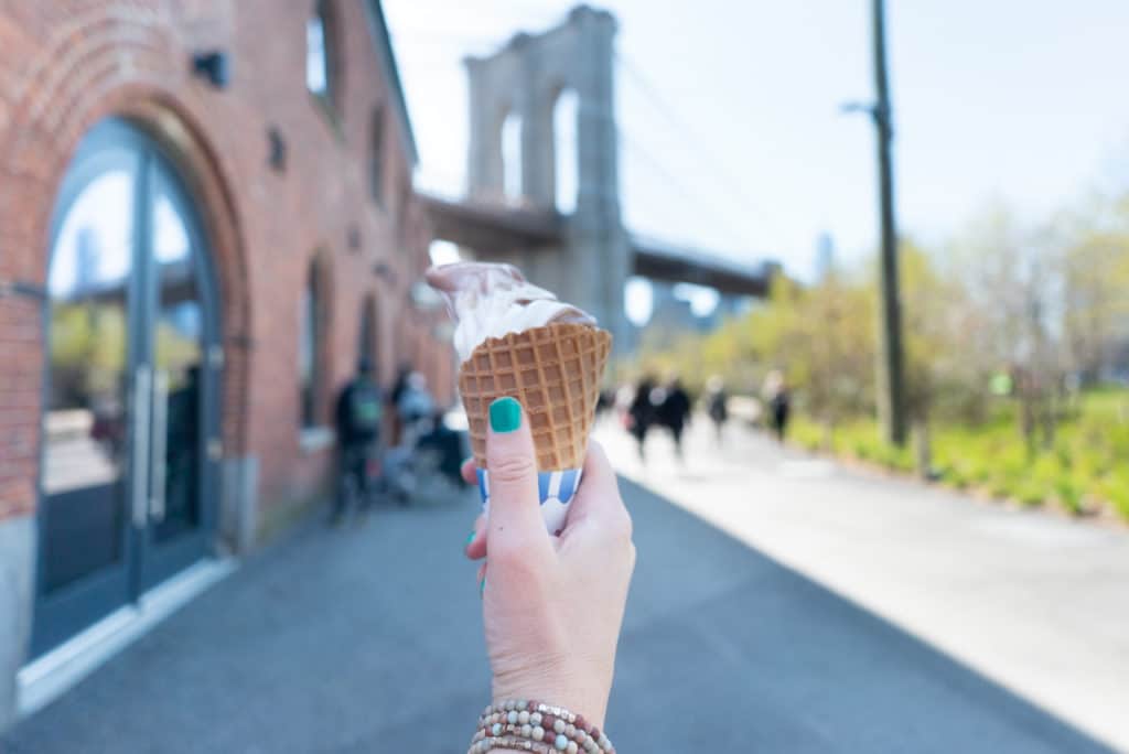 Person's hand holding an ice cream cone with the Brooklyn Bridge in the background. 