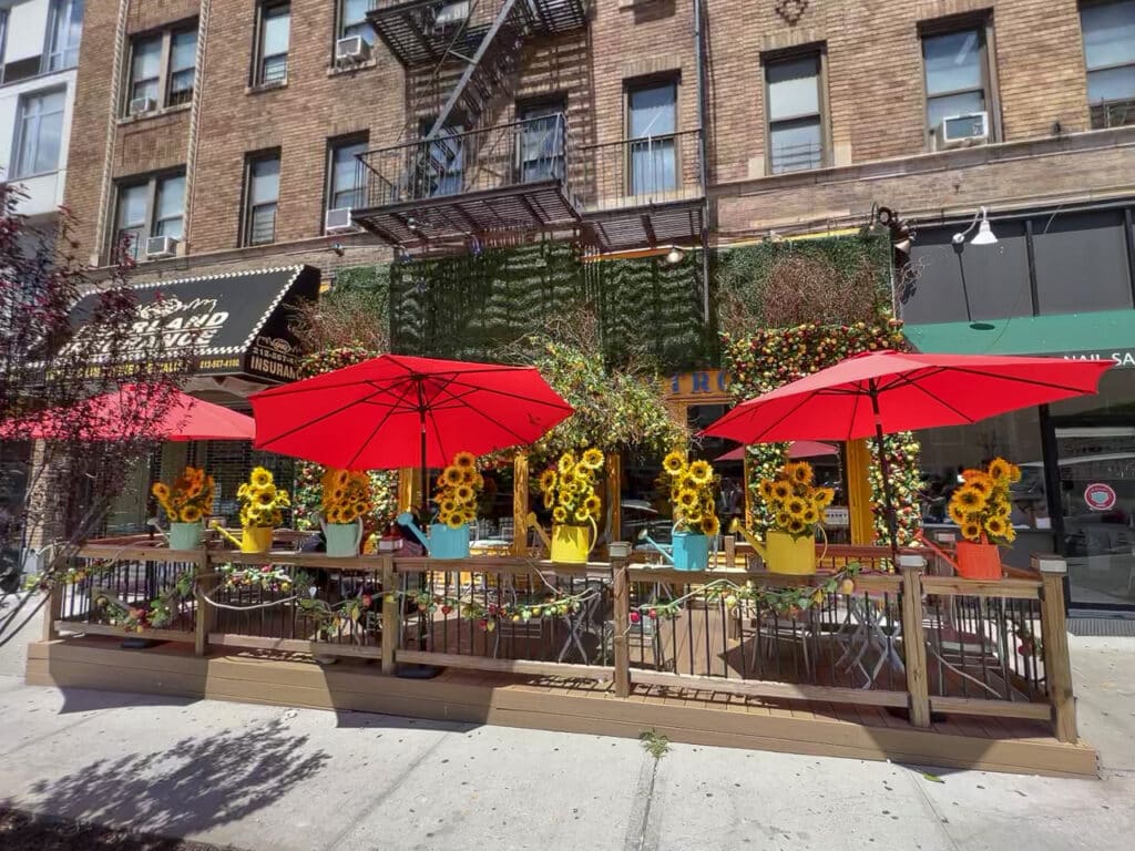 An outdoor dining patio in New York City decorated with yellow sunflowers. Cafe tables are shaded by red umbrellas.