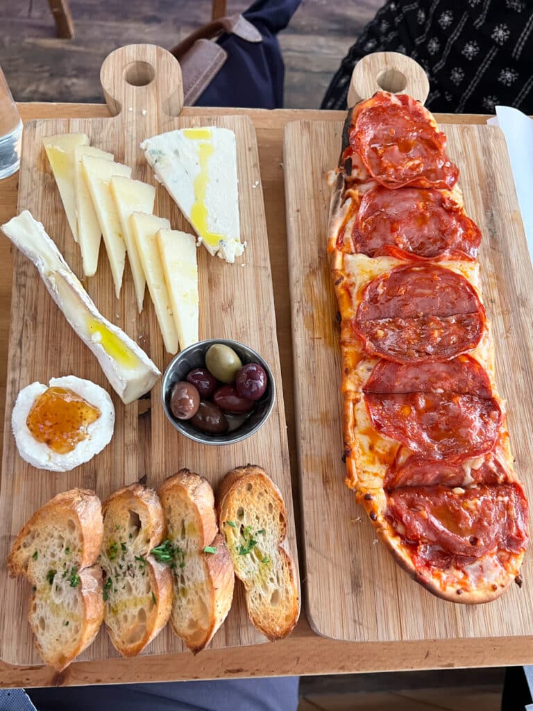 A cheese board with four kinds of cheese, olives, and bruschetta, and a small rectangular pizza, both on wooden serving boards.