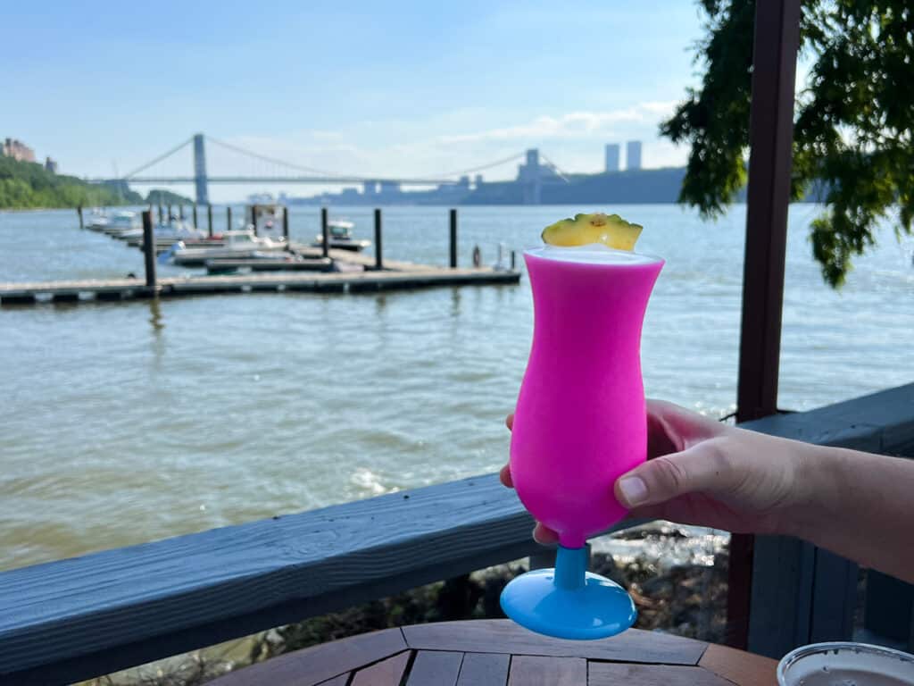 A person's hand holding a frozen cocktail in a bright pink glass with the Hudson River and George Washington Bridge in the background. 