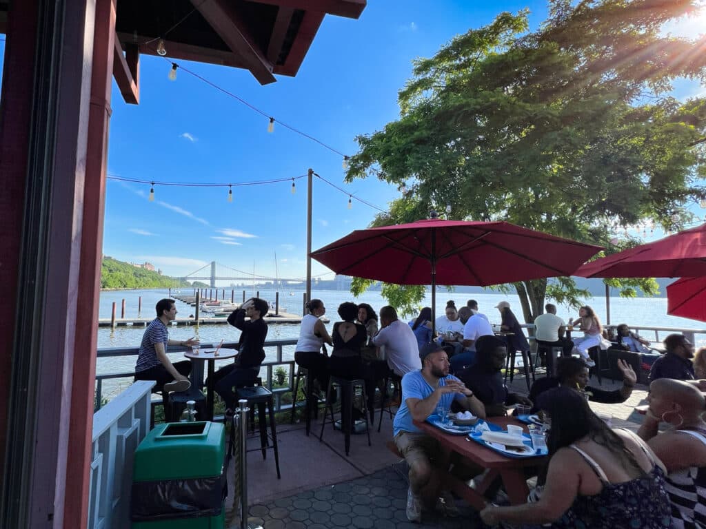 Customers seated at various tables on a patio with views of the Hudson River in the background.