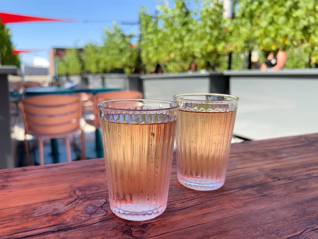 Two glasses of rose wine on a table at a rooftop winery in Brooklyn. Slightly out of focus grape vines are in the background.