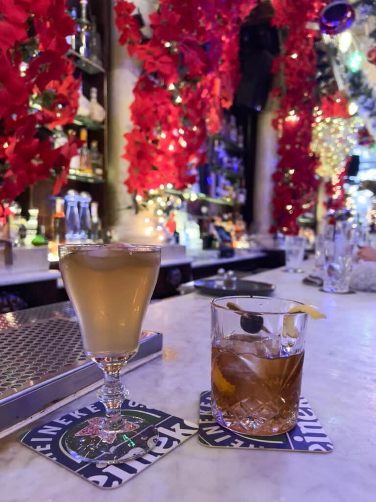 Two cocktails sitting on a bar with Christmas decorations in the background.