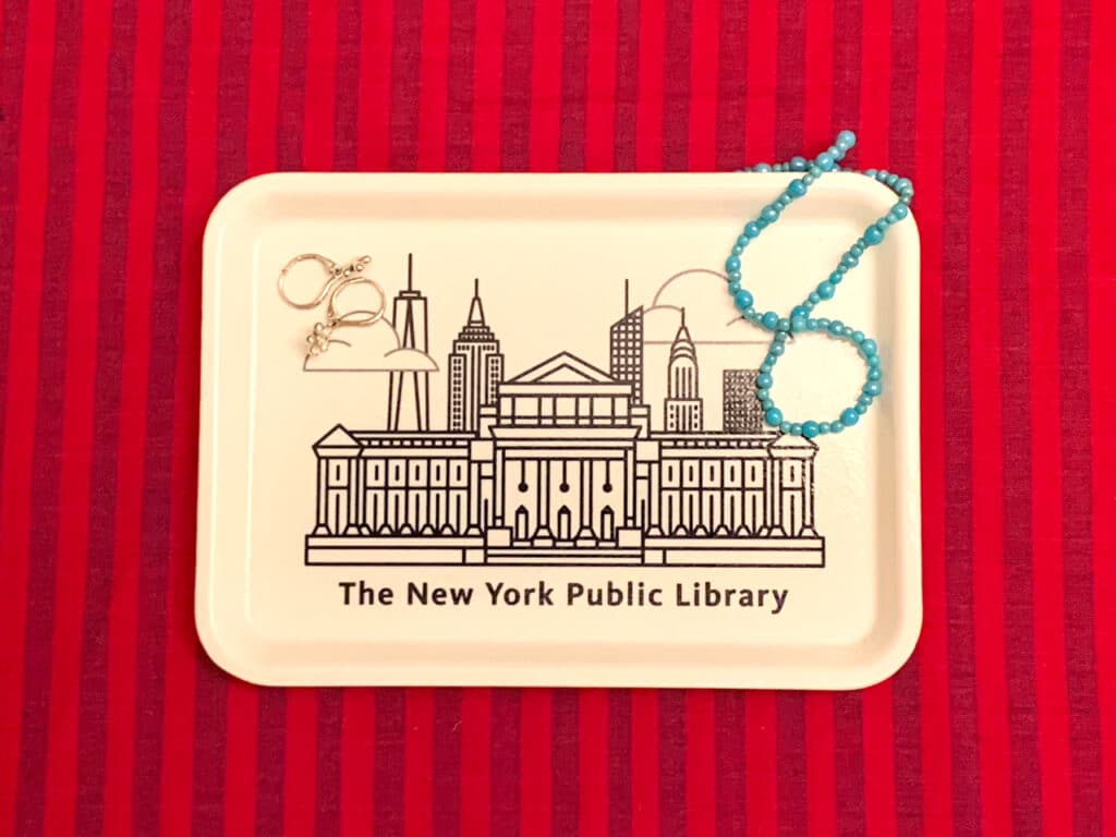 A pair of silver earrings and a blue bead necklace on a white plastic tray with a drawing of the New York Public Library on it. 
