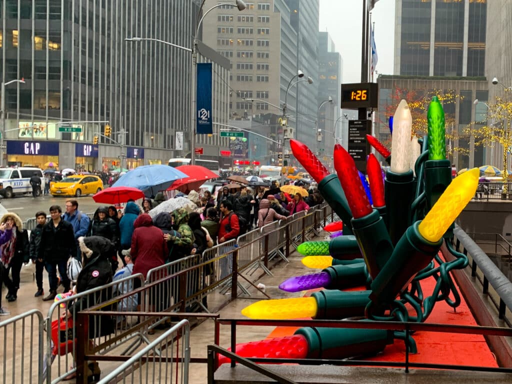 A crowd of people on a rainy, overcast day, walking past Christmas decorations on 7th Avenue in New York City.
