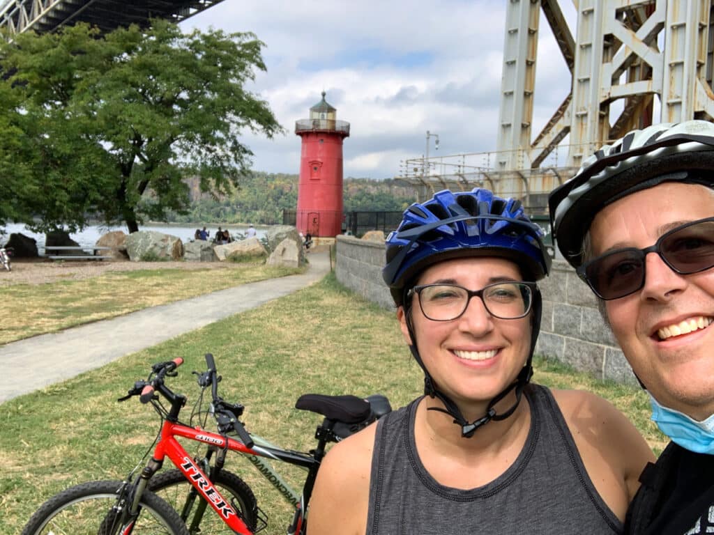 A man and woman wearing bicycle helmets posing in front of their bikes. The Little Red Lighthouse in New York City is in the background.