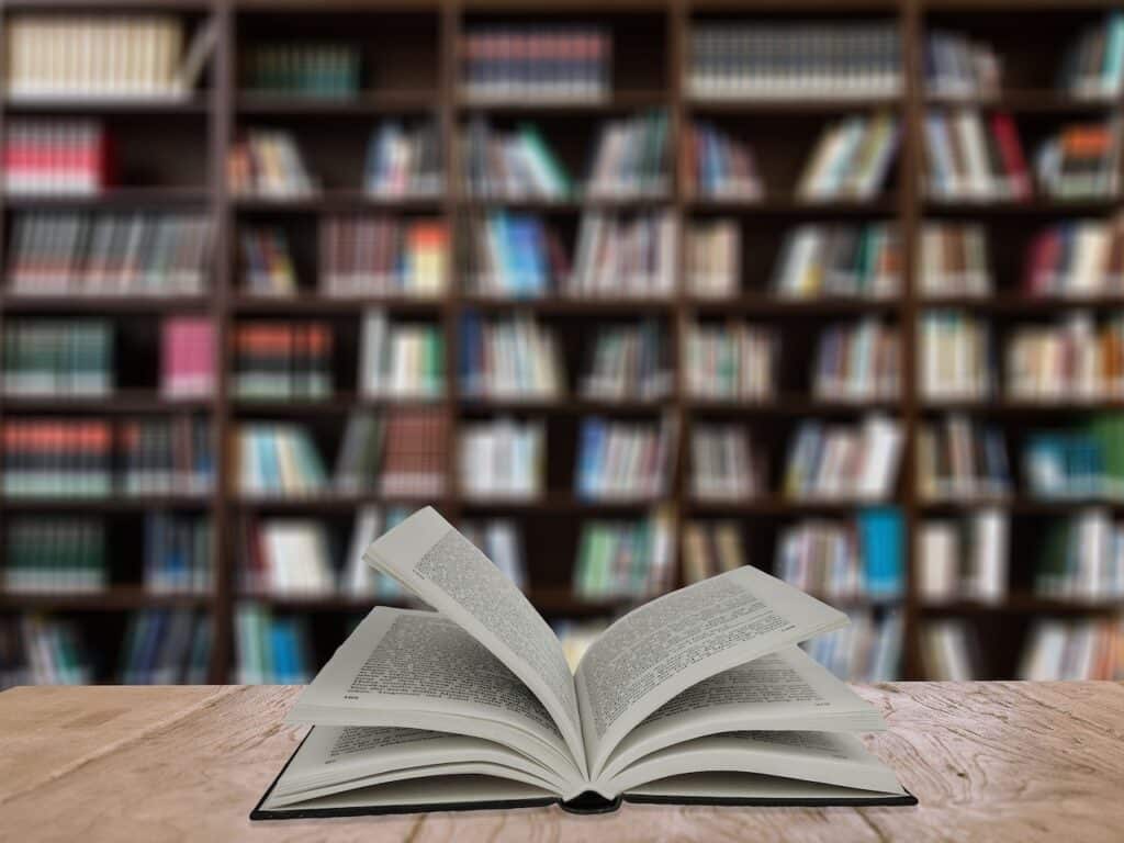 An open book on a table with rows of books on bookshelves, slightly out of focus, in the background.