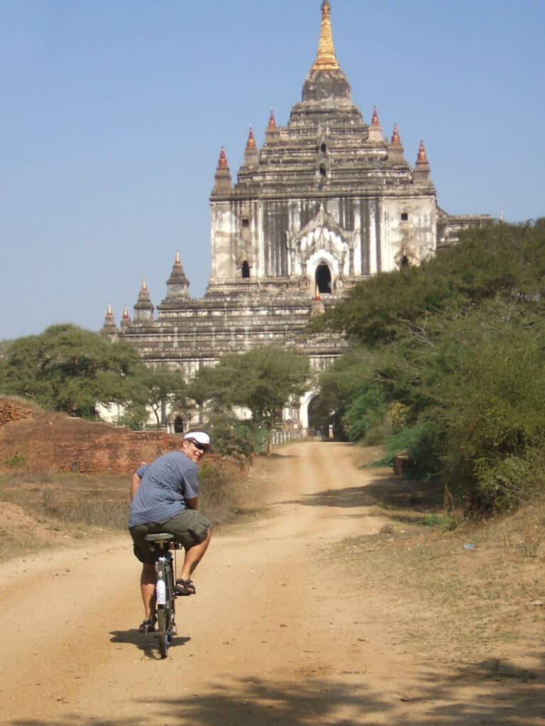 A man riding a bike on a dirt path toward an ancient white stone temple in Bagan, Myanmar.
