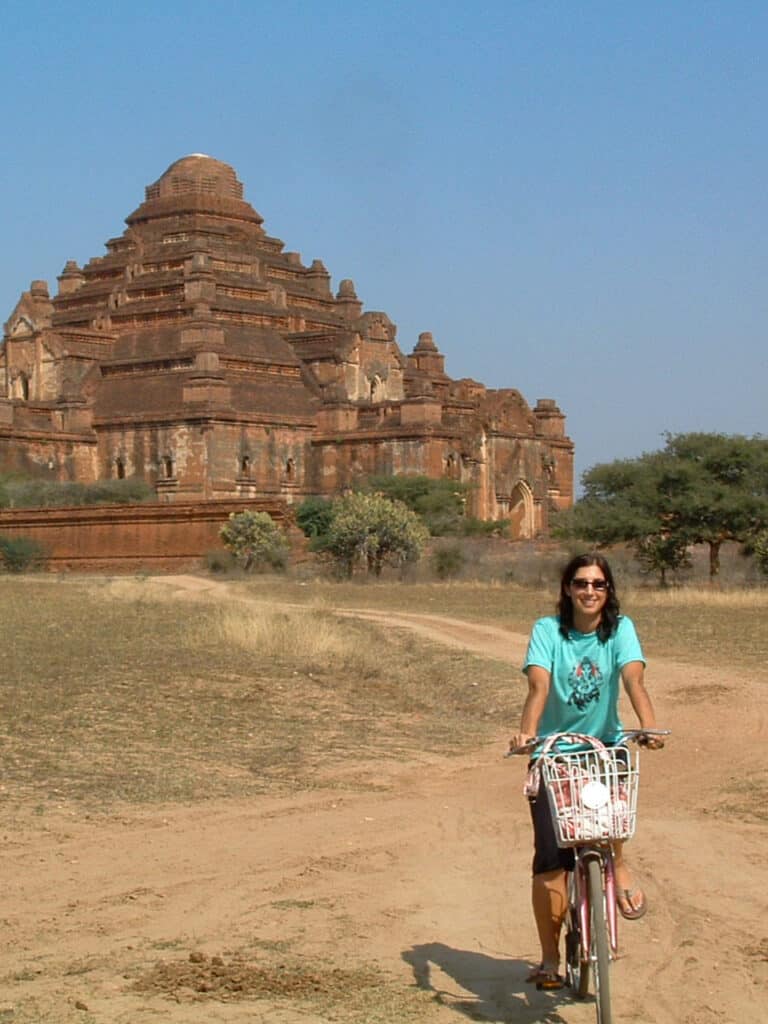 A woman riding a bike on a dirt path in front of an ancient clay-colored stone temple in Bagan, Myanmar.