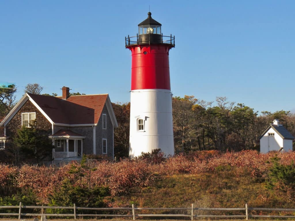 A red and white light house next to a small brown cottage. 