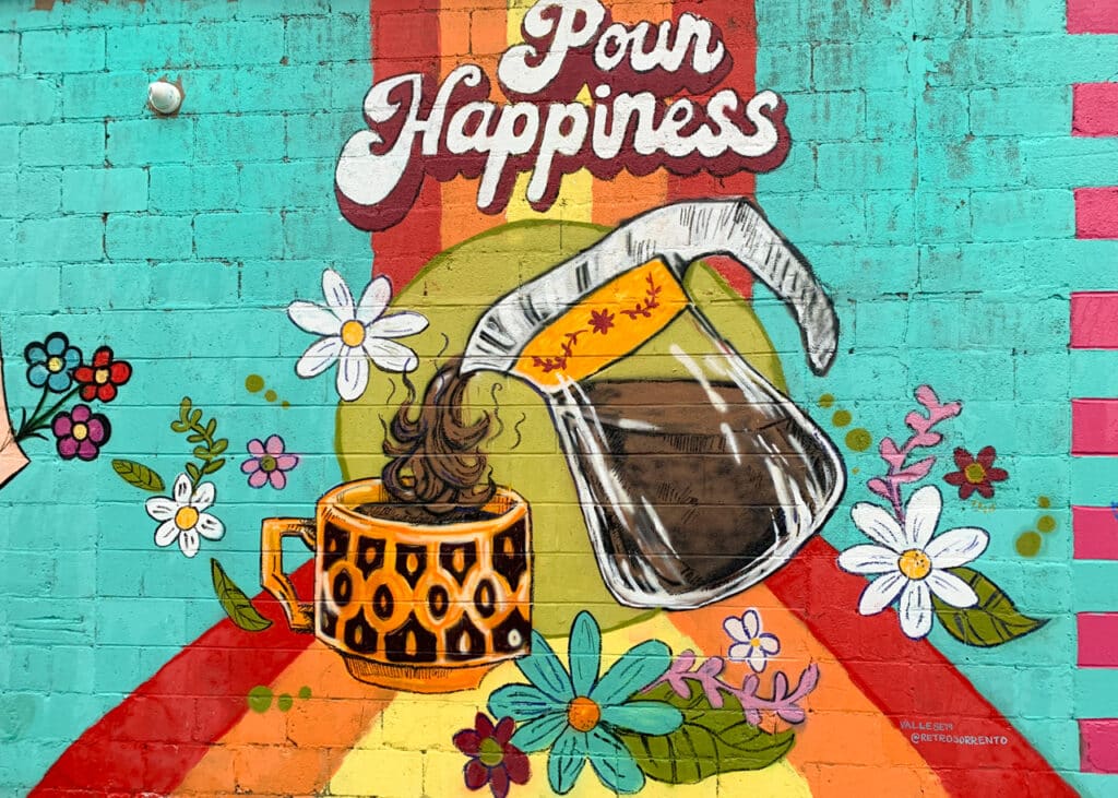 An outdoor wall mural depicting a glass pot pouring coffee into a cup. Pour Happiness is written above the coffee pot.