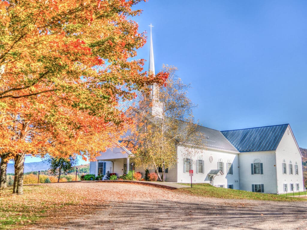 A tree with red and orange leaves in front of a white church, both against the backdrop of a clear blue sky.