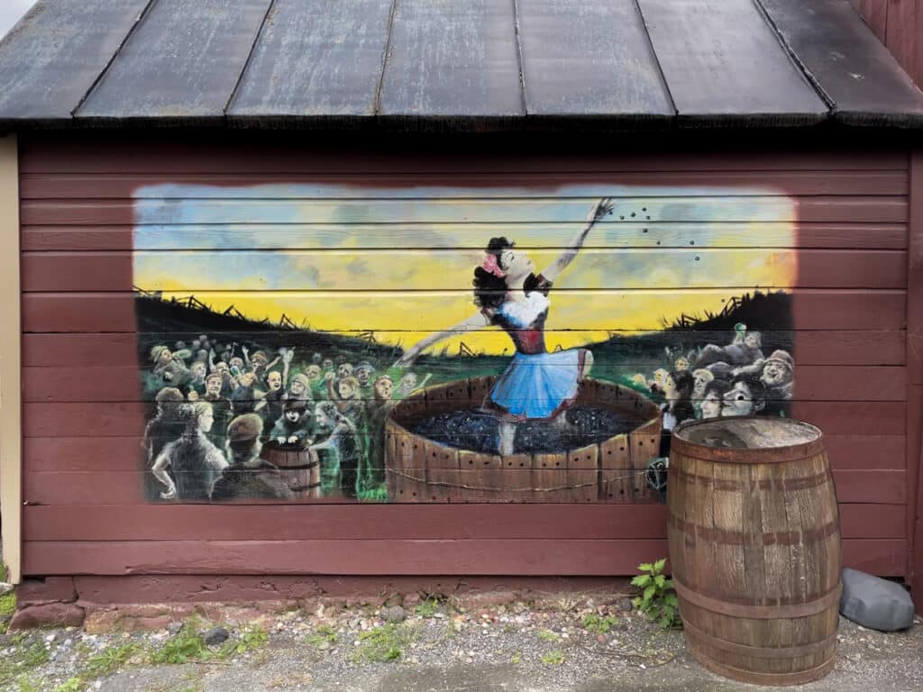 Painting on the side of barn showing a crowd of people watching a woman stomping grapes inside a barrel. 