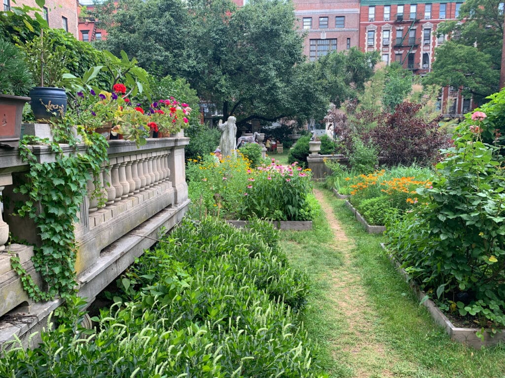 A narrow walking path surrounded by gardens and stone statues at the Elizabeth Street Garden in New York City.
