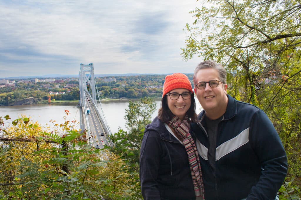 A man and woman posing at the top of a scenic overlook. A bridge over the Hudson River is in the background.