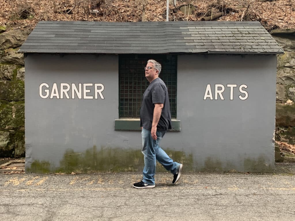 A man walking past a small gray building that has Garner Arts painted on it in white.