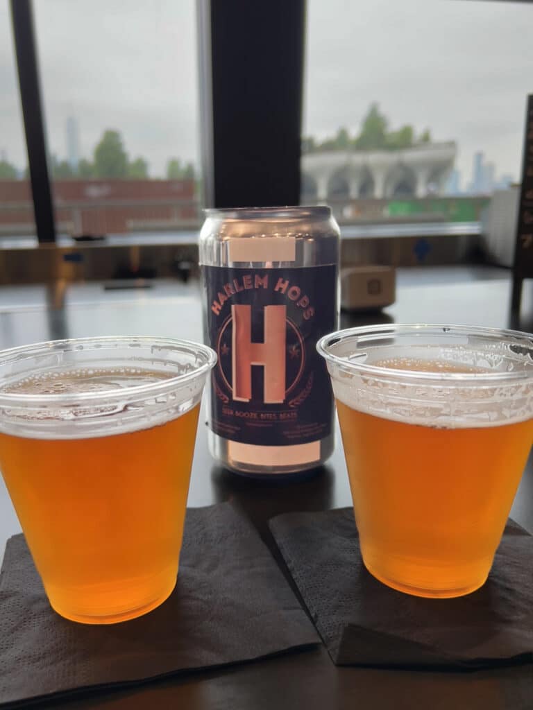 Two beers in plastic cups with a can labeled Harlem Hops behind them. 