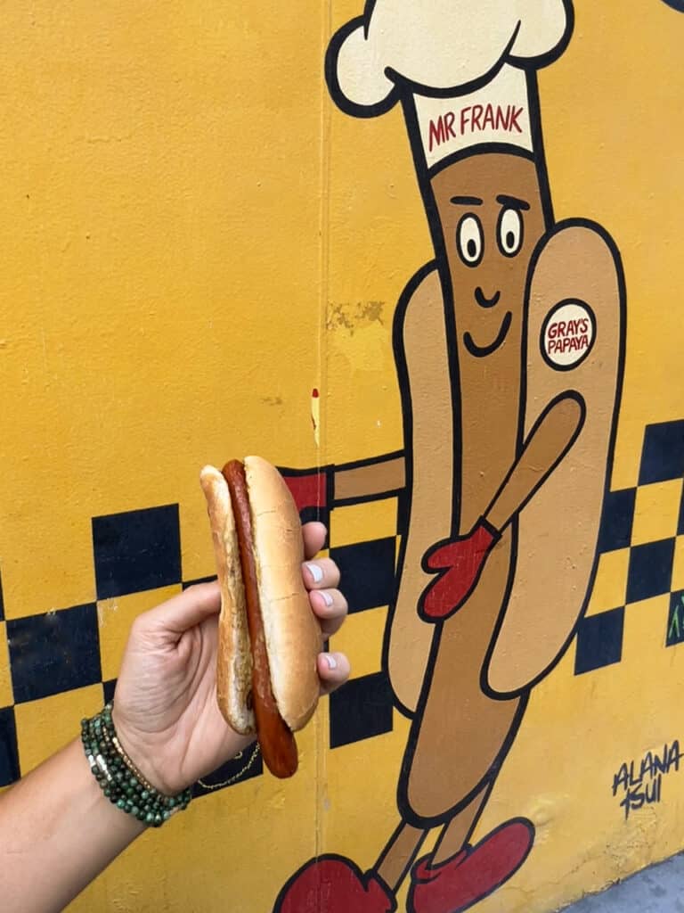 A person's hand holding a hot dog in front of a mural of a hot dog at Gray's Papaya in New York City.