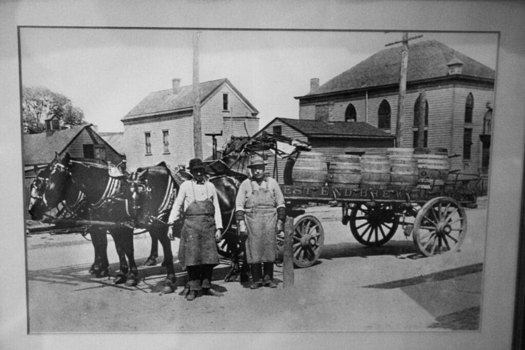 Old black and white photo of two men in aprons standing in front of a horse drawn wagon carrying beer barrels. 