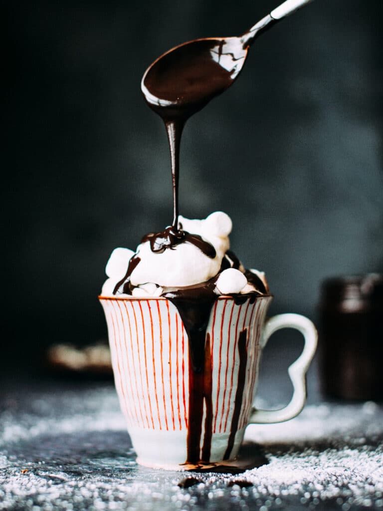 Melted chocolate being drizzled into a cup of hot chocolate topped with whipped cream.