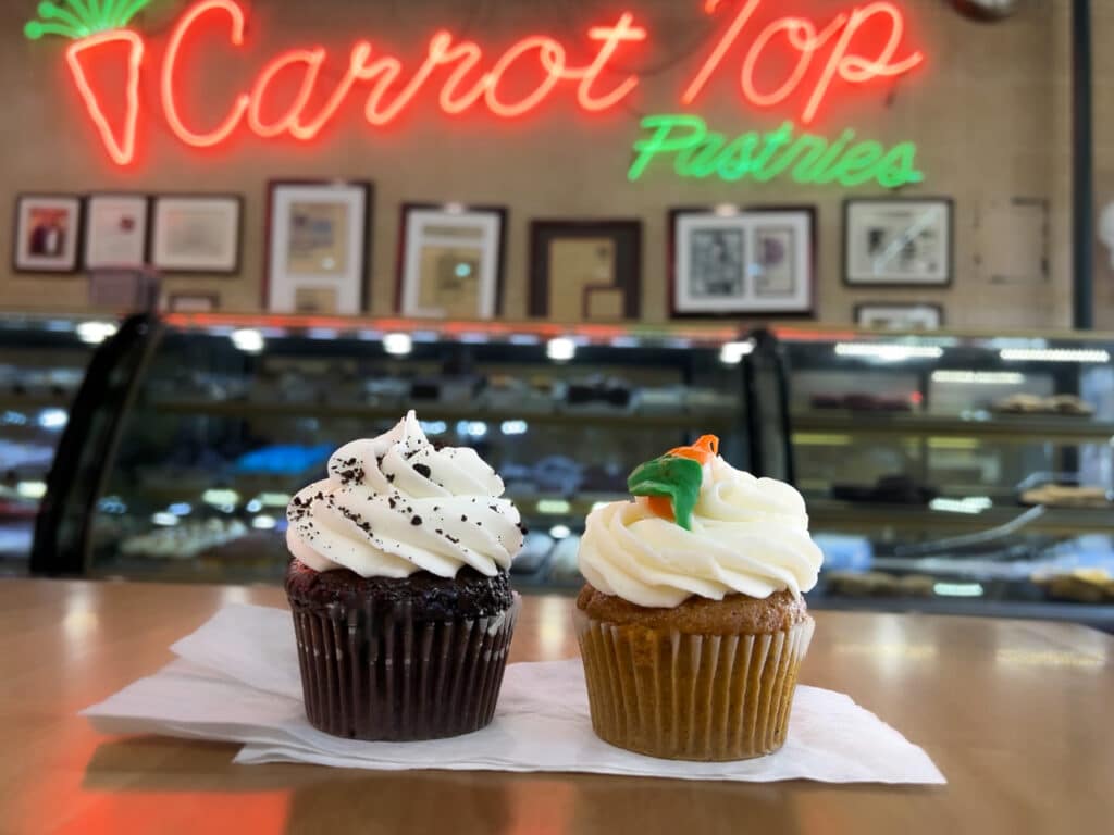 Two cupcakes under a neon sign that says "Carrot Top Pastries."