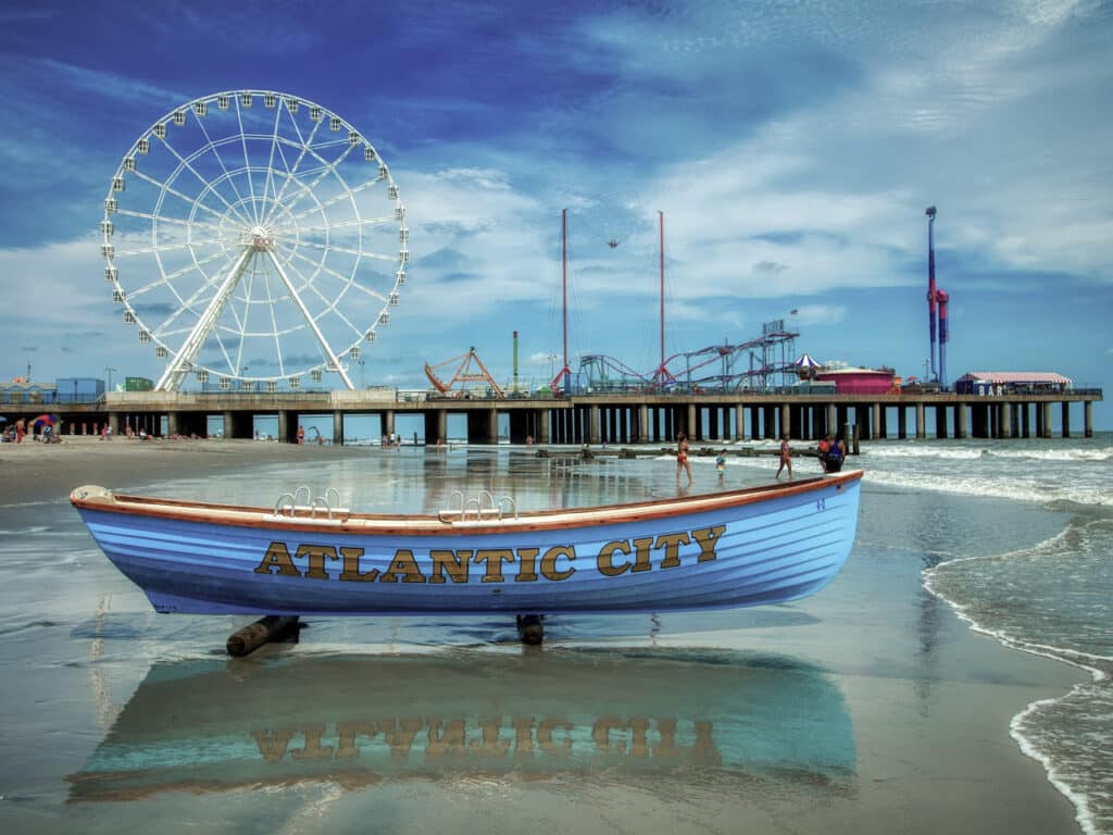 Rowboat that says Atlantic City sitting on the beach, with a pier and ferris wheel in the background.